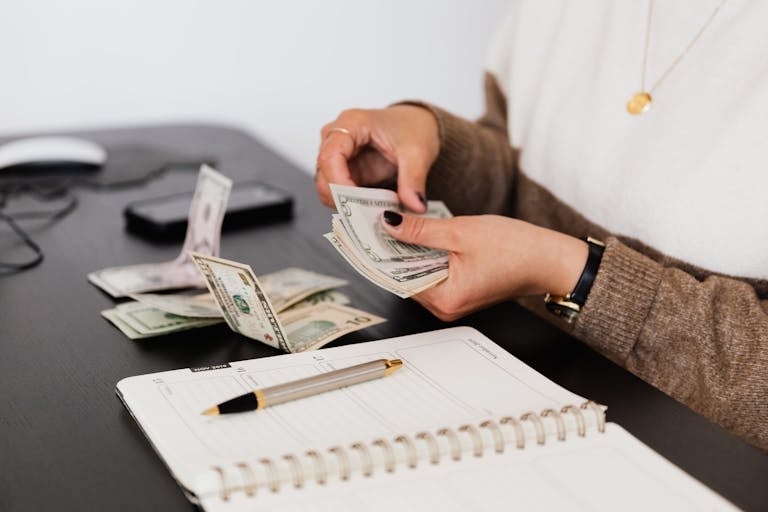 Close-up of person counting cash with Money Hacks budgeting, personal finance, save money, budget plan, financial discipline notepad on desk, indicating financial tasks.